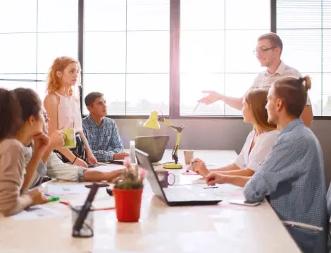 مجموعة من ستة أشخاص في مكتب مشرق ذي نوافذ كبيرة، منخرطون في نقاش حول طاولة عليها أجهزة كمبيوتر محمولة وأوراق. شخص يقف ويتحدث بحماس.